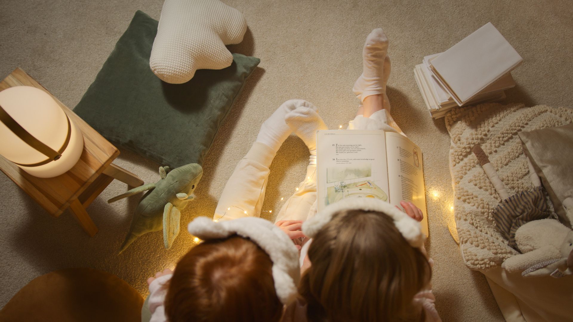 Two young children reading a picture book together on the floor, surrounded by cushions, fairy lights, and soft bedtime textures.